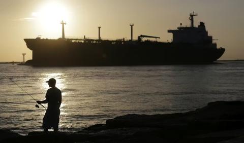 An oil tanker ship with the sun in the background and a man with a cap with a fishing poll in the foreground