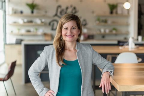 Alison Sizer in a blazer standing in a modern workspace with wooden tables, open shelving, and natural light.