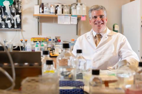A man with silver hair wears a white lab coat, white shirt, and gold tie will sitting behind a lab bench with research equipment on top of it.